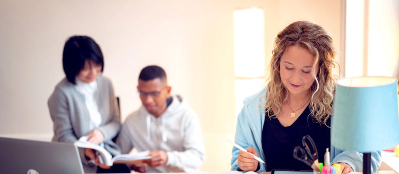 Eine Frau sitzt am Schreibtisch und liest einen Studienbrief. Zwei Studierende sitzen im Hintergrund.